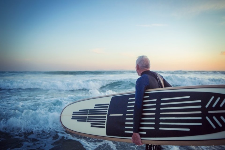 A retiree walking towards the waves at sunset in a wetsuit with a board under his arm